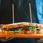 Two stacked sandwich halves on a wooden board with skewers, filled with greens and deli meat.